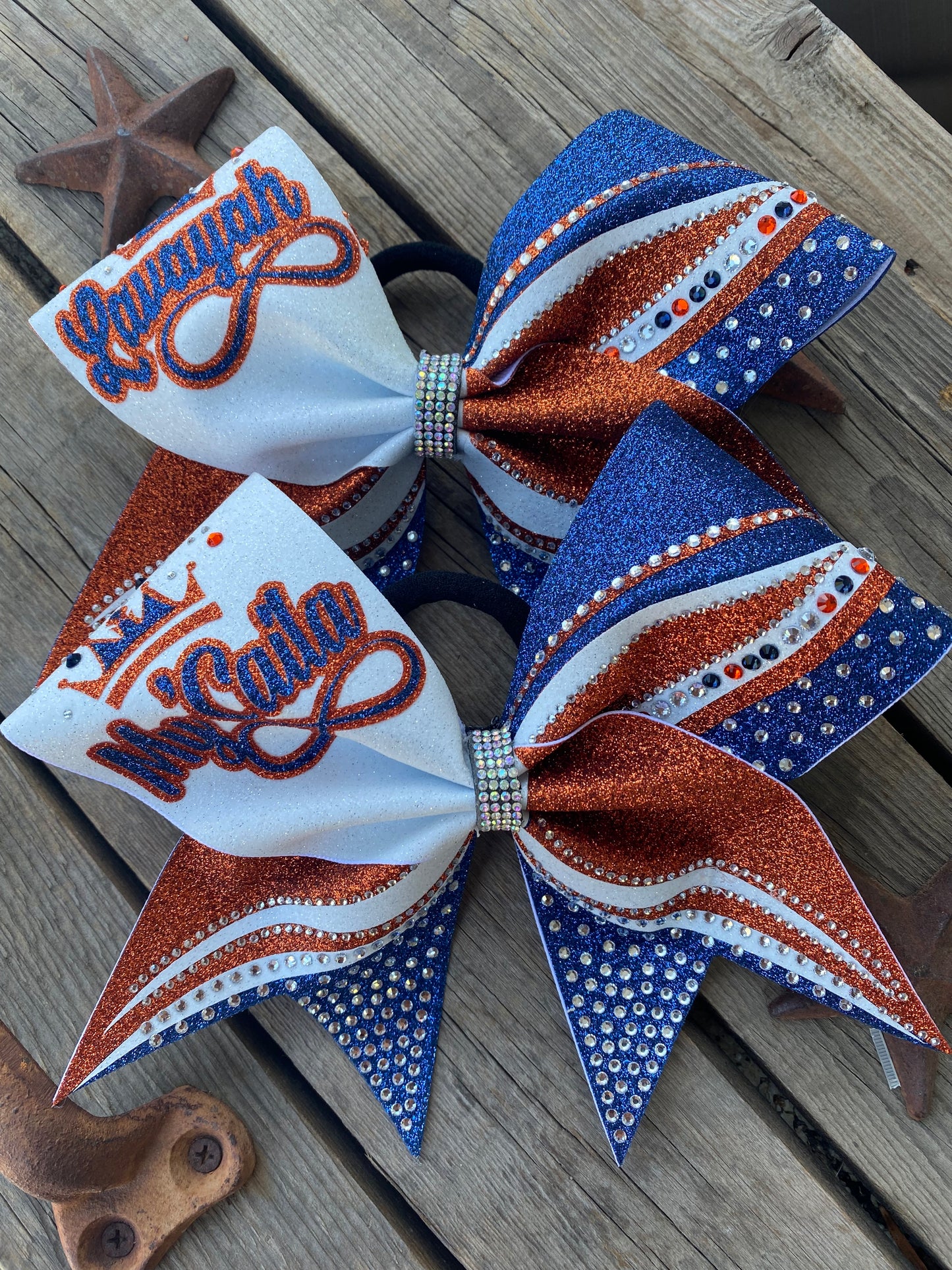 Two cheerleader hair bows on elastic bands, displayed on a weathered wooden surface. The bows feature a white fabric front with orange and blue embroidered lettering, paired with blue glitter, white and orange striped panels, and rhinestone trim.